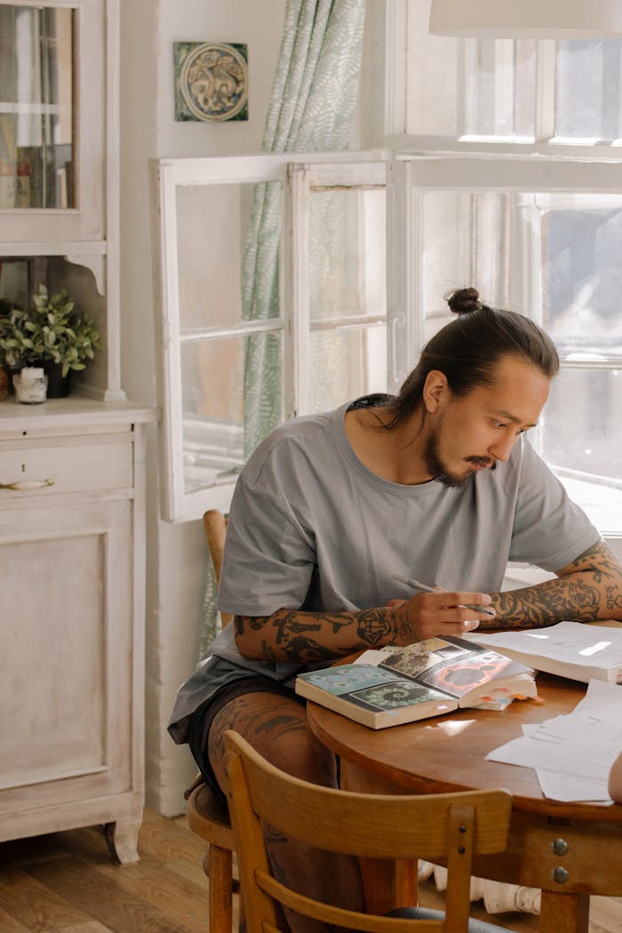 A focused, tattooed man with a bun studies in a cozy room filled with natural light by a window.
