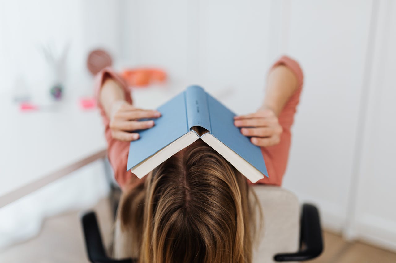 Adult woman covering face with a book in an indoor setting, showcasing study stress.