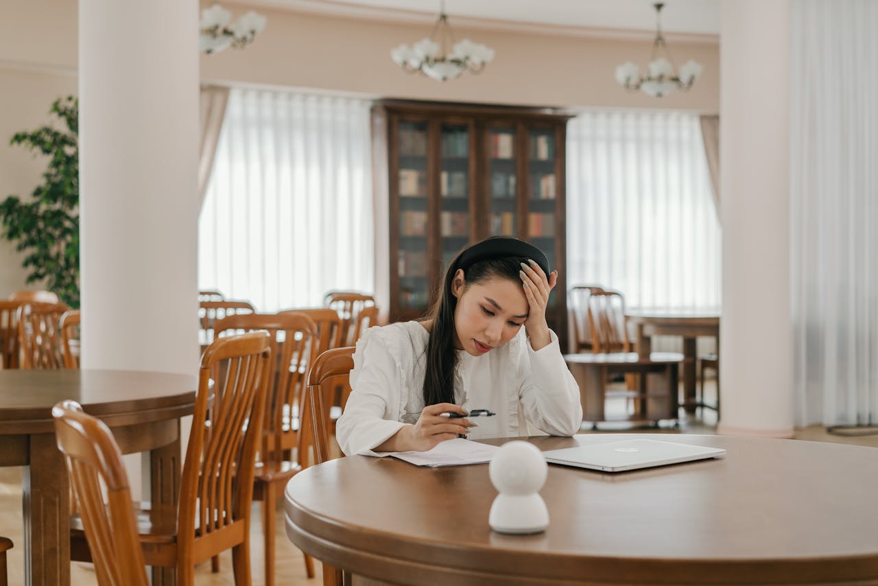 Asian woman focused on study indoors with papers and laptop, reflecting thoughtful concentration.