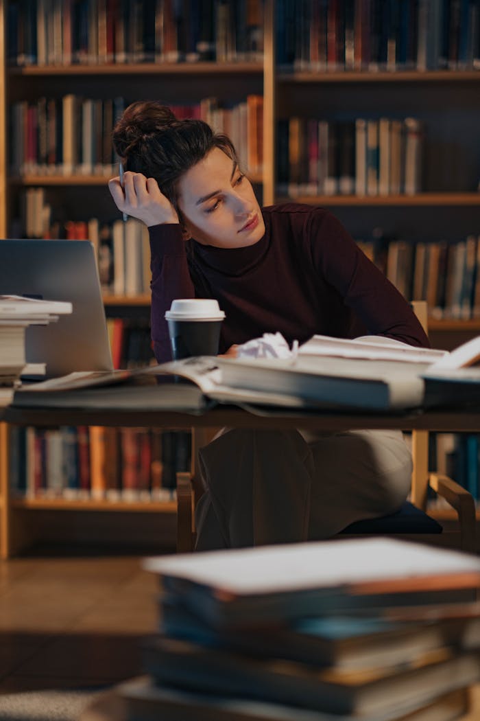 A woman thoughtfully working late in a library filled with books, laptop, and coffee.